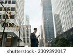Small photo of Young happy Asian businessman executive, Japanese business man investor standing in big city street with downtown buildings using cell phone looking at smartphone checking financial apps on mobile.
