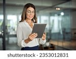Smiling mid aged mature professional business woman banking manager, 40s female executive or entrepreneur holding fintech tab digital tablet standing in office at work, looking at pad.