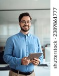 Small photo of Smiling young latin business man manager using tablet computer, happy hispanic businessman employee, company worker looking at camera holding tab working standing in office. Vertical portrait.
