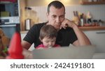 Small photo of father working from home remotely with baby daughter in his arms. pandemic remote work fun business concept. father tries to work at home in kitchen, baby children interfere sitting on their hands