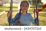 Small photo of school girl swings on swing. concept of a happy childhood and loving family. little child lifestyle in blue dress swings on a swing on the playground, sunset and park in the background