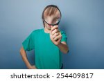 Small photo of boy teenager European appearance brown hair in a shirt looking through a magnifying glass on a gray background, knowledge, foolery