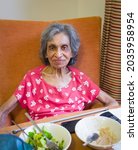 Small photo of Elderly Indian woman in a care home or nursing home, UK, sitting in a chair eating a meal. Depicts frailty and malnutrition in old age