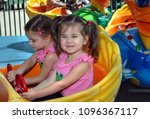 Small photo of Sisters enjoy a kiddie ride at a carnival. One is concentrating on driving, and the other is looking up impishly. Both are wearing pink shirts.