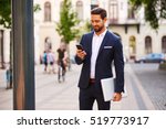 A handsome young businessman holding his laptop while using his phone and standing on the street