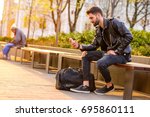 A handsome young hipster man sitting on a bench in the sunset and using his smartphone