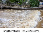 A low view of the river flowing violently past a broken wooden bridge near the bank of an old wooden fence inside a temple in rural Thailand.