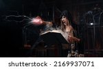 Small photo of Halloween concept. Witch dressed black hood standing dark dungeon room use magic book for conjuring magic spell with lightning. Female necromancer wizard gothic interior put her hand forward screaming
