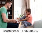 Caucasian girl with braids spending time at home, sitting on a couch while her dad is helping her tying her shoes. Social distancing and self isolation in quarantine lockdown.