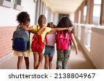 Rear view of multiracial elementary schoolgirls with backpacks and arm around walking in corridor. unaltered, childhood, together, education and back to school concept.