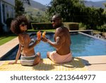 Small photo of Rear view of a couple sitting in the garden on a blanket by a swimming pool wearing beachwear and sunglasses, holding cocktails, turning around and smiling to camera