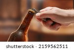 Small photo of Close-up man hand opens lid fresh light beer in sweating from cold dark brown glass bottle with lid opener on blurry background. Light lager is opened with help of bottle opener.