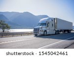 Professional grade big rig semi truck with chrome accessories transporting frozen cargo in refrigerated semi trailer moving on the road along river with bewitching view on the opposite bank