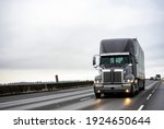 Small photo of Big rig stylish industrial dark gray semi truck with turned on headlights transporting cargo in dry van semi trailer running on the twilight wet road with light reflection surface in rain weather
