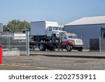 Small photo of Day cab big rig semi towing truck carry another truck with box trailer on flat bed lifted trailer standing on the industrial warehouse parking lot