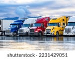 Small photo of Different make big rigs semi trucks tractors with loaded semi trailers standing in the row on truck stop parking lot at early morning waiting for the route continuation time according to the log book