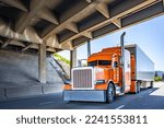 Orange Big rig long haul industrial semi truck tractor with chrome accessories transporting commercial cargo in dry van semi trailer running for delivery on the summer highway road under the bridge