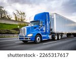 Small photo of Day cab blue big rig industrial semi trucks tractor with roof spoiler and turn on headlight transporting commercial cargo in dry van semi trailer running on the wide highway road at twilight time