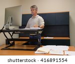 A man is working at a stand up desk in an office where he works because standing is healthier than sitting all day. Live healthy, do not sit all day.