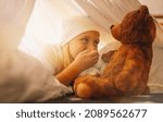 Small photo of Sweet little girl is playing with a teddy bear and holds hand in front of her mouth to keep a secret while lies on her bed in a in tent with nightcap.