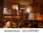 Small photo of man holding wooden bucket and pouring water onto hot stones with rain spoon in sauna room with a group of people. Steam an water on the stones, spa and wellness concept, relax in hot finnish sauna.