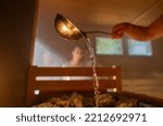 Man pouring water onto hot stone with metal spo in sauna room with a group of people. Steam an water on the stones, spa and wellness concept, relax in hot finnish sauna. Warm temperature bath therapy.