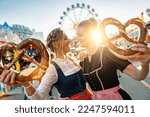 Small photo of girlfriends look to each other with pretzel or brezen on a Bavarian fair or oktoberfest or duld in national costume or Dirndl