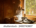 Small photo of Joyful woman in finnish sauna holding ladle with sauna bucket, Finnish hat beside, warm light from window at spa wellness hotel