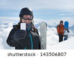 Small photo of Attractive girl holding blank lift pass and ski smiling with snowboarder and mountain range in background. Concept to illustrate ski admission fee
