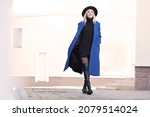 Happy model with beautiful smile walking street in motion. Blonde woman wearing fedora, blue autumn or spring coat, black knitted sweater, shorts, high leather boots. Light background