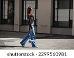 Small photo of Full length beautiful young brunette woman Wearing black leather jacket and wide blue jeans, sneakers, sunglasses and small handbag with chain, walking street on sunny day.