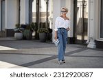 Rejuvenated Trendy mature woman with short hair stands outdoors in city street wears youth clothing white shirt, jeans cargo pants, clutch. Urban style fashion, age and timeless beauty.