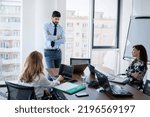 Small photo of Teamwork at the office. Careworn businessman standing at desk while his colleagues sitting at desk and brainstorming.