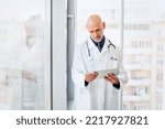 Small photo of Portrait of careworn male doctor holding a digital tablet in his hand while standing on hospital’s foyer.