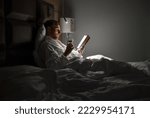 Midle-aged man relaxing rin bed reading book holding a glass of red wine with bedside lamp turned on. Evening relaxation, hobbies, free time concept. Adulthood concept.