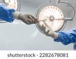Small photo of Nurse sending surgical clamp tool for doctor or surgeon inside operating theatre with medical lamp on background.People did surgery.White wall with space.Person in blue gown with yellow light.