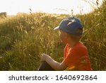 Small photo of LOVING NATURE BOY WEARING BLUE CAP AND ORANGE T SHIRT SITTING IN GREEN GRASS IN SUN LIGHT IN THE SUMMER LANDSCAPE AND LOOKING BACK