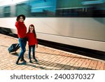 Small photo of Cute sympathetic beautiful smiling young mother with daughter and suitcase on train background on railway station platform