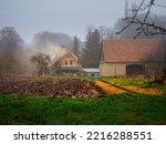 plowed field covered with a snow and old farmhouse on a background in an old village in a rainy misty winter day