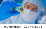Small photo of close up of asian male design engineer in sterile coverall holds microchip with gloves and examines it - ultra modern electronic manufacturing factory