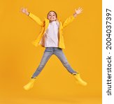 Small photo of Full body of cheerful energetic preteen girl in yellow raincoat and gumboots leaping with outstretched arms and looking at camera against yellow background