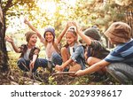 Small photo of Happy kids scouts learning how to create fire with magnifying glass and sun in forest during school camping trip, screaming in excitement while squatting together in nature near pile of dry branches