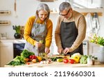 Small photo of Happy elderly couple smiling husband and wife in aprons prepare salad together at kitchen table, chopping variety of colorful vegetables, trying to maintain healthy lifestyle eating vegetarian food