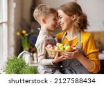 Sweet family portrait of happy mother and little son holding wicker basket full of painted multi-colored Easter eggs, tenderly embracing and smiling in cozy light kitchen at home, selective focus
