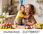 Small photo of Little kid son wearing plush bunny ears kissing and hugging young happy mother while decorating chicken eggs together in kitchen, joyful mom and child preparing for Easter celebration