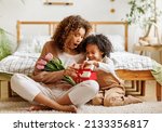 Small photo of Cheerful african american family mom opens a gift box with her son and holding bouquet of flowers while resting on floor by bed during holiday celebration mothers day at home