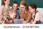 Small photo of Happy female tutor and optimistic children smiling and watching video on tablet while gathering around table during lesson at school