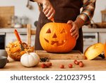 Cropped shot of  man in apron standing in kitchen and carving large orange pumpkin for Halloween party while  making scary face on jack-o-lantern with knife to set the mood for trick-or-treaters 