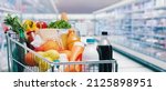 Small photo of Shopping cart filled with food and drinks and supermarket shelves in the background, grocery shopping concept
