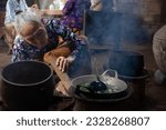 Small photo of Yogyakarta, Indonesia - Feb 12 2023: an old woman cooking on charcoal clay stove smiling in rustic house setting, rather noised and dark mood photo